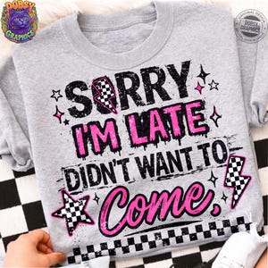 May include: A light gray sweatshirt with the text "SORRY I'M LATE DIDN'T WANT TO COME" in black and pink. The design includes stars and checkered lightning bolts. The sweatshirt has a crew neck and long sleeves.