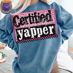 May include: A blue sweatshirt with the words "Certified Yapper" in a black and white design with pink accents. The design is on the back of the shirt. The sweatshirt has a crew neck and long sleeves.