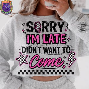 May include: A light gray sweatshirt with the text "SORRY I'M LATE DIDN'T WANT TO COME" in black and pink. The design includes stars, lightning bolts, and a checkered pattern. The sweatshirt has long sleeves.