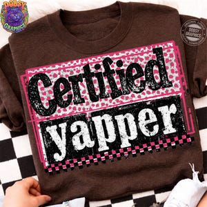 May include: A brown sweatshirt with the words "Certified Yapper" in a black and white distressed font. The text is set against a pink and white heart pattern and a black and pink checkered border.