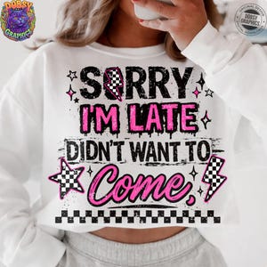 May include: White sweatshirt with the text "SORRY I'M LATE DIDN'T WANT TO Come" in black and pink. The design includes stars, a checkered lightning bolt, and a checkered star. The sweatshirt has long sleeves.