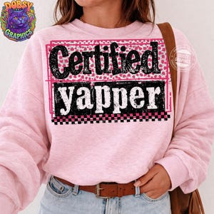 May include: A light pink sweatshirt with the words "Certified Yapper" in a black, pink, and white design. The text is in a bold, playful font, with a leopard print background. The sweatshirt has long sleeves.