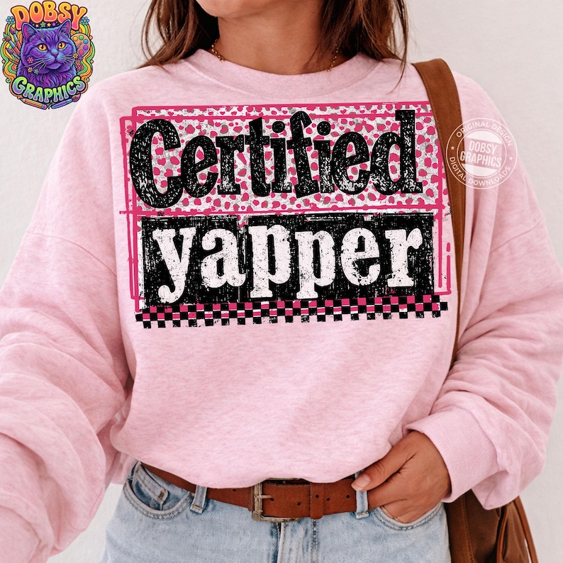 May include: A light pink sweatshirt with the words "Certified Yapper" in a black, pink, and white design. The text is in a bold, playful font, with a leopard print background. The sweatshirt has long sleeves.