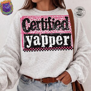 May include: A light gray sweatshirt with the words "Certified Yapper" in a black, pink, and white design. The top text is in a pink heart pattern, and the bottom text is in a black and white checkered pattern.