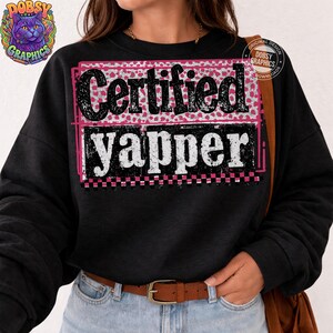 May include: Black sweatshirt with the text "Certified Yapper" in a white, pink, and black design. The text is inside a pink and white patterned rectangle. The sweatshirt has a crew neck and long sleeves.