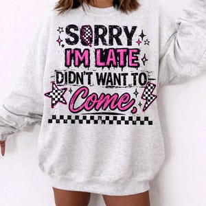 May include: A light gray sweatshirt with the text "SORRY I'M LATE DIDN'T WANT TO COME" in black and pink. The design includes stars, a checkered flag, and a lightning bolt. The bottom of the sweatshirt has a checkered pattern.