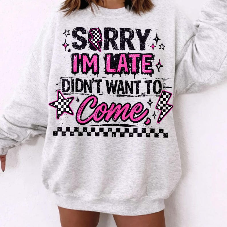 May include: A light gray sweatshirt with the text "SORRY I'M LATE DIDN'T WANT TO COME" in black and pink. The design includes stars, a checkered flag, and a lightning bolt. The bottom of the sweatshirt has a checkered pattern.