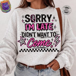 May include: A light gray sweatshirt with the text "SORRY I'M LATE DIDN'T WANT TO COME" in black and pink. The design includes stars, a checkered flag, and lightning bolts. A brown leather belt is visible.