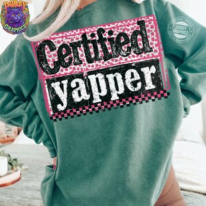 May include: A teal sweatshirt with the words "Certified Yapper" in a black and white design. The text is set against a pink and white polka dot background, with a pink border and a checkered pattern at the bottom.