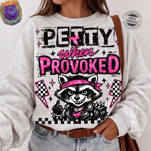 May include: A light gray sweatshirt with the text "PETTY when PROVOKED" in black and pink. The graphic features a raccoon wearing a pink bandana and a black leather jacket, giving the middle finger. The design includes lightning bolts and stars.