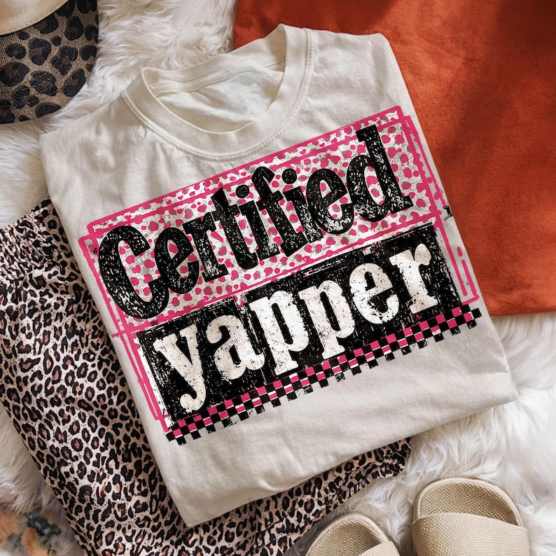 May include: Cream-colored t-shirt with the text "Certified Yapper" in a pink and black design. The shirt is laid on a white fur-like surface, with leopard print pants and a hat nearby. A pair of beige sandals are also visible.