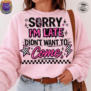 May include: A light pink sweatshirt with the text "SORRY I'M LATE DIDN'T WANT TO COME" in black and pink. The design includes stars, a checkered flag, and a lightning bolt. The sweatshirt has long sleeves.
