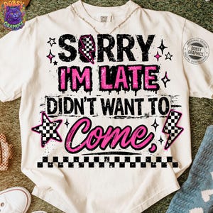 May include: Cream-colored t-shirt with the text "SORRY I'M LATE DIDN'T WANT TO COME" in black and pink. The design includes stars, a checkered lightning bolt, and a checkered border at the bottom. A necklace is visible.