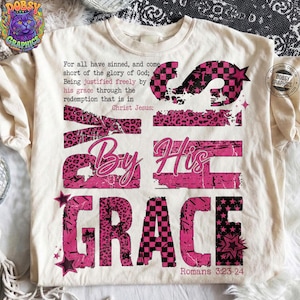 May include: Cream-colored t-shirt with a pink and black graphic design. The design includes the words "S By His GRACE" in various fonts and patterns, along with text from Romans 3:23-24.