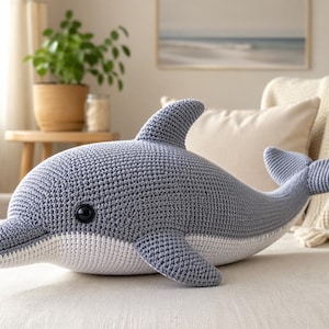 以下が含まれることがあります： グレーと白のグラデーションの、かぎ針編みで作られたイルカのぬいぐるみ。イルカは、ひれ、尾、黒い目の笑顔の顔が特徴です。ぬいぐるみはクリーム色の表面に置かれています。