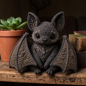 Puede incluir: Una figura de murciélago de ganchillo en tonos grises y marrones, con grandes ojos negros y alas detalladas. Tiene un pequeño colgante de cristal y está colocada sobre una estantería de madera. Una maceta con una suculenta y libros antiguos en el fondo.