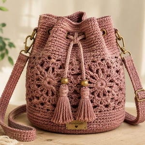 May include: A dusty rose crochet bucket bag featuring an intricate floral pattern, a drawstring closure with two tassels, wooden beads, and a rectangular metal tag labeled Handmade with a heart icon. It has an adjustable strap with brass hardware.