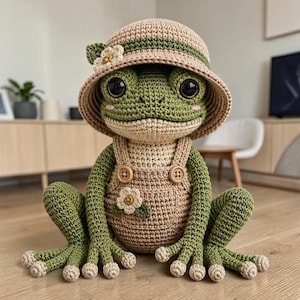 Peut inclure: Une peluche grenouille crochetée verte portant un chapeau bob beige avec une bande verte et une petite fleur blanche. La grenouille porte une salopette crochetée beige avec deux boutons en bois et une fleur blanche appliquée sur le devant.