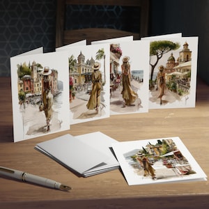 May include: A set of folded greeting cards featuring watercolor illustrations of individuals in sun hats walking through European coastal towns. Each card displays a unique scene with architectural details, greenery, and a signature in the corner.