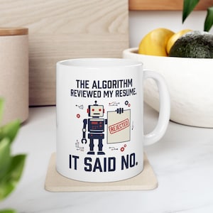 Puede incluir: Taza de cerámica blanca con un robot que sostiene un letrero de "RECHAZADO". La taza tiene el texto "THE ALGORITHM REVIEWED MY RESUME. IT SAID NO." en negro y rojo. La taza está sobre un posavasos beige.