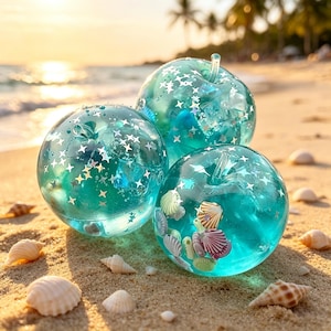 Puede incluir: Tres adornos transparentes en forma de manzana llenos de líquido azul, estrellas plateadas y conchas marinas. Los adornos están sobre una playa de arena con conchas marinas esparcidas. El fondo muestra el océano y una puesta de sol.