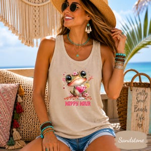 May include: A beige tank top featuring a graphic of a cartoon frog holding a pink cocktail with the text Hoppy Hour printed in pink below. The frog is decorated with small pink flowers and the design includes pink splatters around the illustration.
