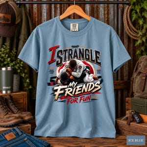 May include: A light blue t-shirt featuring a graphic of two individuals in white martial arts uniforms engaged in a grappling match. The text reads I STRANGLE MY FRIENDS FOR FUN, with additional small text labels for specific grappling techniques.