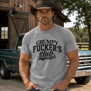 May include: A heather grey short-sleeved t-shirt featuring black text that reads GRUMPY FUCKER'S club LIFETIME MEMBER. The shirt has a crew neckline and is worn with a brown cowboy hat. The design is centred on the chest in a bold, vintage-style font.