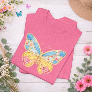 以下が含まれることがあります： ピンク色のTシャツ。カラフルな蝶のデザインが特徴です。蝶の羽はパッチワークで、青、黄色、ピンクのセクションがあり、花柄とギンガムチェックのパターンが組み込まれています。Tシャツは白い木の表面に折りたたまれており、背景には花と帽子があります。