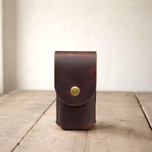 May include: A dark brown leather pouch with a vertical rectangular shape and a rounded flap closure. The pouch is secured by a single circular brass-coloured metal snap button centred on the front flap, featuring visible stitching along the sides.