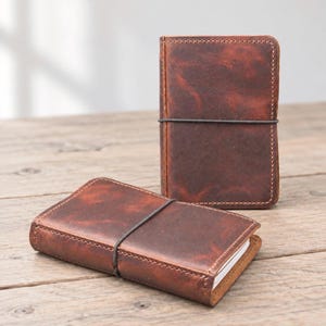 May include: Two rectangular notebooks with dark brown, distressed leather covers. Each notebook is secured by a thin, black elastic band wrapped vertically around the centre. The leather features visible grain and tonal variations in a deep, rustic brown.