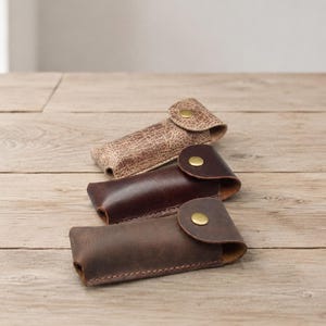 Peut inclure: Trois étuis rectangulaires en cuir avec des rabats arrondis et des boutons-pression couleur laiton. L'étui du haut est beige clair avec une surface texturée craquelée, celui du milieu est marron foncé lisse et celui du bas est en cuir marron mat.