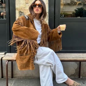 Könnte beinhalten: Eine braune Wildlederjacke mit langen Fransen an den Ärmeln und Schultern, getragen über einem schlichten weißen T-Shirt und einer locker sitzenden weißen Hose. Das Outfit wird mit flachen, spitz zulaufenden Leoparden-Ballerinas und einer braunen Schildpatt-Sonnenbrille kombiniert.