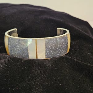 May include: A silver cuff bracelet with a central section of speckled gray and white segments. The bracelet has a gold-colored border and a central vertical stripe. The bracelet is on a black surface.