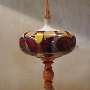 Könnte beinhalten: Handgefertigtes dekoratives Holzobjekt mit glänzendem Finish. Das Stück hat einen bauchigen, gemusterten Mittelteil mit roten, gelben und braunen Akzenten, der von einem gedrehten Sockel getragen wird. Die Oberseite hat eine schlanke, spitze Spitze.
