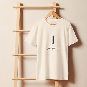 Puede incluir: Una camiseta de manga corta y cuello redondo en color blanco roto, con el texto azul marino oscuro J for jacquère impreso en el centro de la parte delantera. La prenda se muestra en una percha de madera sobre una pared de tono claro.