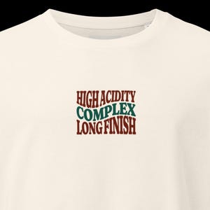 Puede incluir: Una camiseta de color crema con un gráfico central que contiene el texto HIGH ACIDITY en rojo oscuro, COMPLEX en verde oscuro y LONG FINISH en rojo oscuro. Las letras tienen un aspecto ondulado y distorsionado en el centro del pecho.
