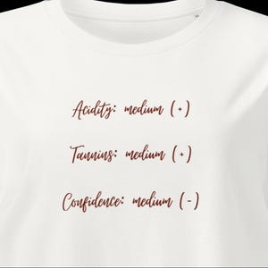 May include: A white t-shirt featuring dark red cursive text that reads: Acidity: medium (+), Tannins: medium (+), and Confidence: medium (-). The shirt has a round neckline and short sleeves, presenting a clean, simple design with clear lettering.
