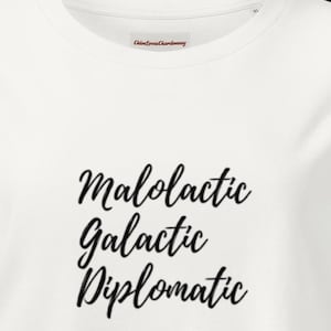 Puede incluir: Una camiseta blanca con el texto negro Malolactic, Galactic, Diplomatic impreso en una fuente cursiva en el pecho. La camiseta tiene un cuello redondo y una pequeña etiqueta interior con el texto ChloeLovesChardonnay y la talla XL.