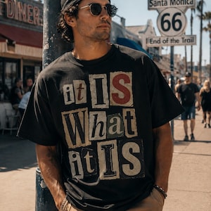 Peut inclure: Un t-shirt noir à manches courtes arborant un imprimé graphique avec le texte "it IS What it IS" dans un style lettre anonyme avec des arrière-plans rectangulaires variés. Le t-shirt a un col rond et une coupe décontractée en tissu sombre.