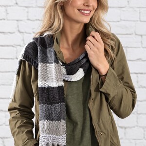 May include: A knitted scarf featuring a horizontal striped pattern in shades of black, charcoal grey, light grey, and white. The scarf is constructed with a textured knit stitch and finished with long, matching fringe tassels at the ends.