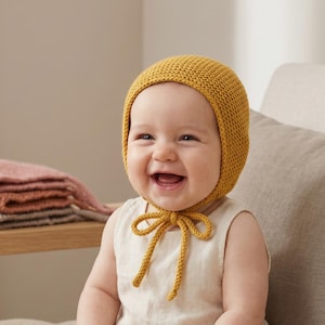 Puede incluir: Un bebé sonriente con un gorro de punto amarillo mostaza con una cinta a juego. El bebé también lleva un body de color claro. Una sonaja de madera y mantas dobladas están en el fondo.