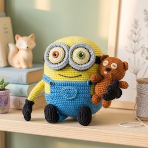 May include: A crocheted Minion doll in yellow and blue dungarees, holding a small brown teddy bear. The Minion has large, round eyes with grey frames. The doll is on a wooden shelf with other decorative items.
