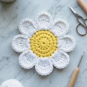 Puede incluir: Un posavasos de margarita hecho a mano con ganchillo, con pétalos blancos y un centro amarillo. El posavasos está rodeado de herramientas de artesanía, incluyendo unas tijeras, ganchillos y una madeja de hilo blanco. El posavasos tiene un diámetro de unos 15 cm.