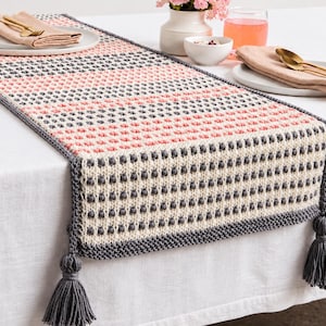 Puede incluir: Un camino de mesa rectangular de ganchillo con un patrón de puntos bobble texturizados en rayas horizontales alternas de color crema, rosa suave y gris oscuro. El camino tiene un borde gris oscuro y borlas a juego en cada esquina.