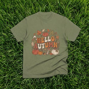 Può includere: Una t-shirt a maniche corte color verde oliva con una grafica centrale che riporta il testo "HELLO AUTUMN" in un carattere grassetto e arrotondato. Il design include illustrazioni di zucche arancioni e bianche, vari funghi e foglie autunnali.