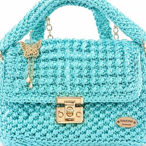Puede incluir: Un bolso de mano de ganchillo turquesa con asa superior y correa de cadena dorada. El bolso presenta un cierre de broche dorado y un dije de mariposa dorada decorativa. La etiqueta dice "RAMONA handmade".
