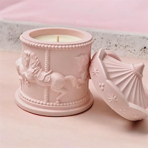 Puede incluir: Un tarro de vela rosa claro con tapa a juego, que presenta un diseño de caballo de carrusel en relieve en el exterior. El recipiente cilíndrico tiene un borde con cuentas y una mecha central, mientras que la tapa cónica está decorada con estrellas.