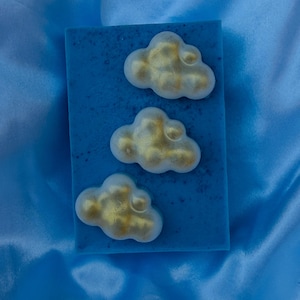 Pode incluir: Uma barra retangular de sabonete azul com textura salpicada. A superfície é decorada com três adornos em relevo em forma de nuvem branca, cada um com um acabamento dourado metálico no topo, dispostos diagonalmente sobre a base azul.