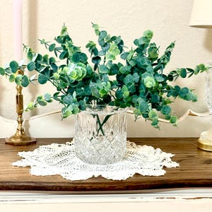 May include: A clear glass vase with a diamond-cut pattern containing a cluster of artificial eucalyptus stems. The stems feature numerous small, rounded, sage and dark green leaves that extend outward in a dense, multi-directional arrangement.